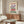 May include: Vintage-style poster with cream background featuring purple irises and red flowers, bold text reading "FLOWER MARKET" and "AMSTERDAM" displayed above wooden credenza with brass hardware. Modern white curved chair and glass-top coffee table visible in minimalist living room with beige walls and natural lighting.