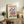 May include: Vintage-style travel poster featuring purple iris flowers and pink blooms with "Flower Market Amsterdam" text, displayed in light wood frame on rustic wooden sideboard alongside fern plant, ceramic vases, and books in cozy living room with beige armchair and neutral walls