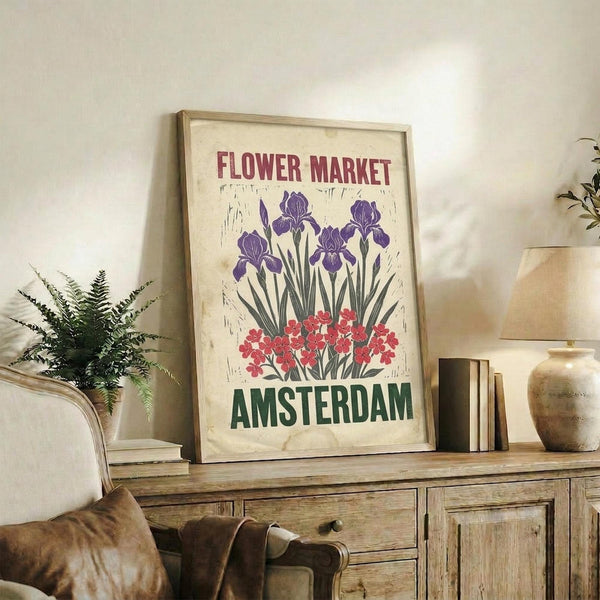 May include: Vintage-style travel poster featuring purple iris flowers and pink blooms with "Flower Market Amsterdam" text, displayed in light wood frame on rustic wooden sideboard alongside fern plant, ceramic vases, and books in cozy living room with beige armchair and neutral walls