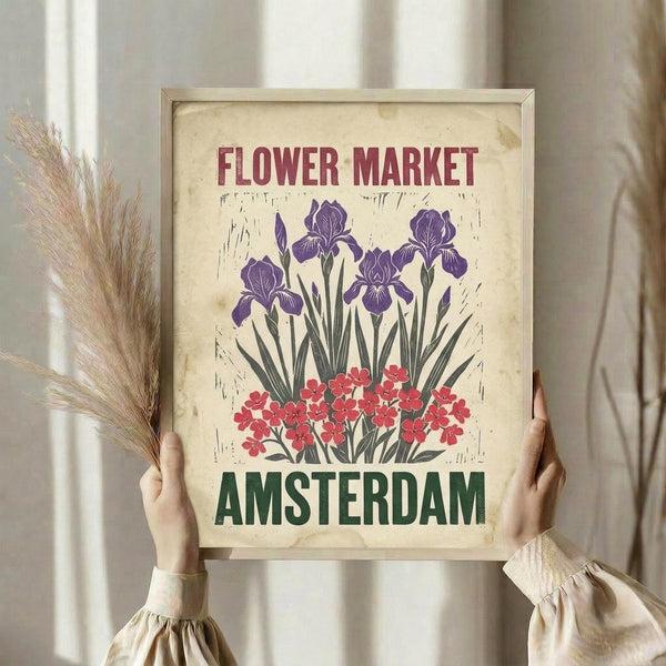 May include: Person in beige clothing holding framed vintage-style poster featuring purple irises and red flowers on aged cream background with "FLOWER MARKET" and "AMSTERDAM" text. Dried wheat stems visible to left, soft natural lighting from window creates warm atmosphere in neutral-toned room setting.