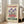 May include: A vintage-style poster with cream background featuring purple iris flowers and red blooms, with "FLOWER MARKET" in red text at top and "AMSTERDAM" in green text at bottom, displayed in a light wooden frame leaning against a white wall next to a beige upholstered sofa in a bright living room with large windows and natural lighting.