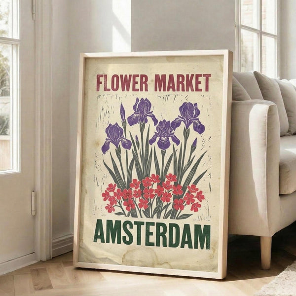 May include: A vintage-style poster with cream background featuring purple iris flowers and red blooms, with "FLOWER MARKET" in red text at top and "AMSTERDAM" in green text at bottom, displayed in a light wooden frame leaning against a white wall next to a beige upholstered sofa in a bright living room with large windows and natural lighting.