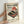 May include: Hands holding a framed vintage-style Japanese poster featuring a red turntable with cherry blossom branches, a red sun, and bonsai tree against cream background. Black Japanese text at top, wooden frame, displayed against white wall.