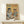 May include: Framed vintage-style poster with yellow background showing two anthropomorphic cats at cafe table - tabby cat in beret reading "Cat Tales" newspaper with coffee cup, orange cat in sunglasses and floral shirt laughing. Table has checkered cloth, pastries, coffee pot. Small wrapped gift sits on light wood shelf below frame against white wall.