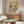 May include: Vintage-style poster showing two anthropomorphic cats at a breakfast table - one tabby in beret reading newspaper, one orange cat in sunglasses and Hawaiian shirt holding coffee cup. Yellow background with "CAT CAFÉ" text in blue and red letters. Framed artwork displayed above mid-century modern walnut credenza in bright living room with white curved chair, glass coffee table, and natural lighting.