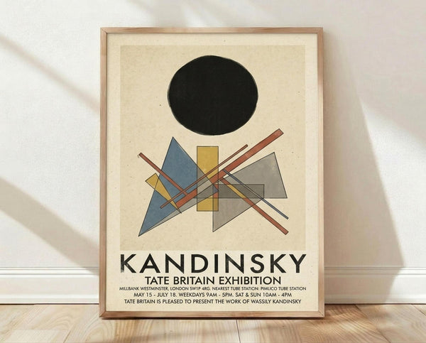 May include: Framed exhibition poster leaning against white wall on light wooden floor. Features abstract geometric composition with black circle, blue triangle, yellow rectangles, gray shapes, and intersecting red and white lines on cream background. Bold "KANDINSKY" text below with Tate Britain exhibition details. Natural wood frame with soft lighting and fabric shadows visible.