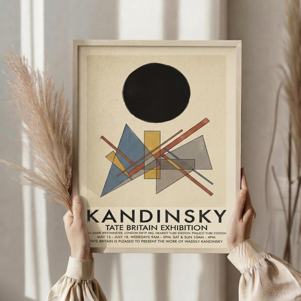 May include: Person in cream sweater holding framed Kandinsky exhibition poster featuring black circle above geometric shapes in blue, yellow, orange and gray on cream background. Natural light setting with dried pampas grass and white curtains visible.