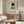 May include: Kandinsky exhibition poster with black circle and geometric shapes in blue, yellow, and red displayed above wooden sideboard. White modernist chair and glass coffee table in minimalist room with beige walls and natural lighting.