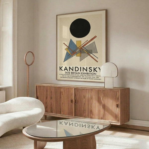 May include: Kandinsky exhibition poster with black circle and geometric shapes in blue, yellow, and red displayed above wooden sideboard. White modernist chair and glass coffee table in minimalist room with beige walls and natural lighting.