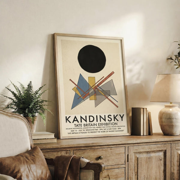May include: Framed Kandinsky exhibition poster with black circle and geometric shapes in blue, yellow, and red tones, displayed on rustic wooden sideboard beside fern plant, ceramic vase, and stacked books in neutral-toned living room with leather armchair