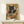 May include: Framed poster print displayed on light wood shelf against white wall, showing black cat with golden eyes eating ramen noodles from decorative bowl with chopsticks, Japanese text and geometric patchwork background in earth tones, small wrapped gift box with greenery sits beside frame