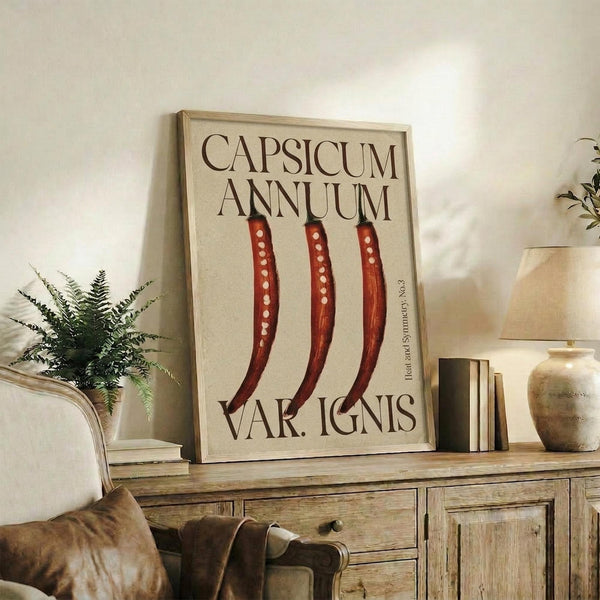 May include: Framed botanical poster displaying three red chili peppers with white spots against cream background, featuring Latin text. Shown leaning against white wall on rustic wooden sideboard with drawers, surrounded by fern plant, stacked books, ceramic vase, and brown leather chair in cozy living room setting with warm natural lighting.