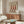 May include: Modern living room interior featuring botanical wall art displaying three red chili peppers with white seeds against cream background, hung above mid-century walnut credenza. Scene includes white curved chair, brass floor lamp, round glass coffee table with colorful abstract art, and natural lighting creating shadows on neutral walls.