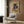 May include: Framed poster showing black cat with yellow eyes eating ramen noodles from decorated bowl, Japanese text and colorful geometric background in vintage style. Modern living room setting with beige armchair, brass floor lamp, dark wood paneled wall, and patterned throw pillow.