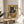 May include: Framed poster print featuring a black cat with golden eyes eating ramen noodles with chopsticks, surrounded by Japanese text and geometric patterns in warm yellow, green, and orange tones. The artwork is displayed on a light wood round side table next to a beige upholstered chair, with natural sunlight streaming across white walls and a potted plant visible in the corner.