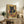 May include: Framed poster displaying a black cat with golden eyes eating ramen noodles from a red bowl, rendered in vintage Japanese art style with geometric patterns in warm earth tones. Print sits on rustic wooden credenza alongside books, ceramic vase, and fern plant in cozy living room setting with beige armchair and neutral walls.