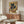 May include: Framed poster displaying a black cat with yellow eyes eating ramen noodles with chopsticks, featuring Japanese text and colorful geometric background in earth tones. Shown in modern living room with wooden credenza, white curved chair, glass coffee table, and warm natural lighting.