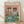 May include: Framed poster print leaning against white wall on light wood floor. Shows two stylish cats wearing sunglasses at dining table - one in Hawaiian shirt and straw hat, another in striped shirt with red bow tie. Pink background with teal table displaying wine glasses, fish on plate, tuna cans, kibble bowls, and "Catnip Brew" bottle. Colorful illustrated art style in natural wood frame.