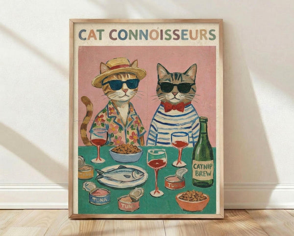 May include: Framed poster print leaning against white wall on light wood floor. Shows two stylish cats wearing sunglasses at dining table - one in Hawaiian shirt and straw hat, another in striped shirt with red bow tie. Pink background with teal table displaying wine glasses, fish on plate, tuna cans, kibble bowls, and "Catnip Brew" bottle. Colorful illustrated art style in natural wood frame.