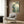 May include: Framed Japanese landscape print featuring mountain valley with pine trees in blue-green tones, displayed on wood paneled wall above modern beige armchair with geometric pillow, brass floor lamp and dark side table with open book nearby