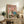 May include: Framed poster with pink background showing white duck in chef's hat and person in denim jacket holding cooked fish, with prepared dishes and wine bottles below. Displayed on rustic wooden sideboard with fern plant, books, and ceramic vase in modern living room setting with beige chair visible.