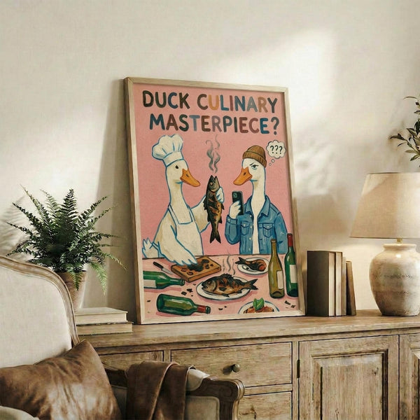 May include: Framed poster with pink background showing white duck in chef's hat and person in denim jacket holding cooked fish, with prepared dishes and wine bottles below. Displayed on rustic wooden sideboard with fern plant, books, and ceramic vase in modern living room setting with beige chair visible.