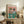 May include: Framed poster displaying two illustrated cats dressed as food connoisseurs - one wearing a straw hat and floral shirt, the other in striped shirt with bow tie, both with sunglasses. They sit behind a table with wine glasses, fish, bowls of food, and a "Catnip Brew" bottle on pink and teal background. Print is propped on rustic wooden sideboard next to books, plant, and ceramic vase in cozy living room setting.