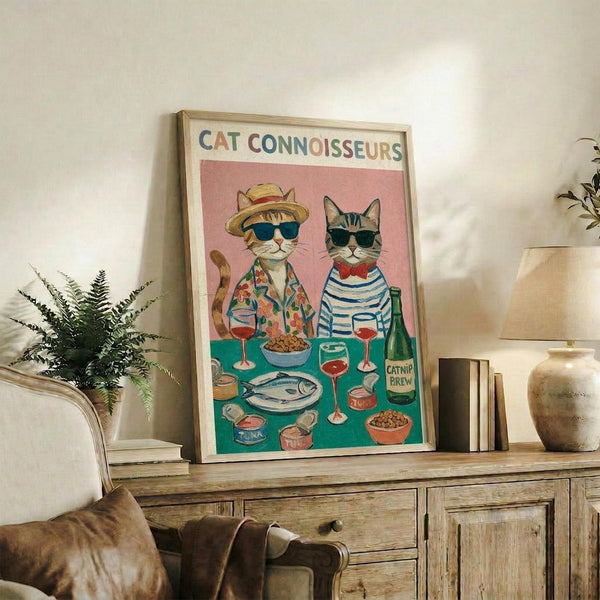 May include: Framed poster displaying two illustrated cats dressed as food connoisseurs - one wearing a straw hat and floral shirt, the other in striped shirt with bow tie, both with sunglasses. They sit behind a table with wine glasses, fish, bowls of food, and a "Catnip Brew" bottle on pink and teal background. Print is propped on rustic wooden sideboard next to books, plant, and ceramic vase in cozy living room setting.