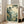 May include: Framed vintage Japanese woodblock print artwork leaning against white wall next to beige upholstered chair. The print features misty mountains, tall pine trees, and traditional houses in a valley, rendered in muted greens, blues, and cream tones. Light wooden frame surrounds the serene landscape scene. Natural sunlight streams through nearby windows onto hardwood flooring.