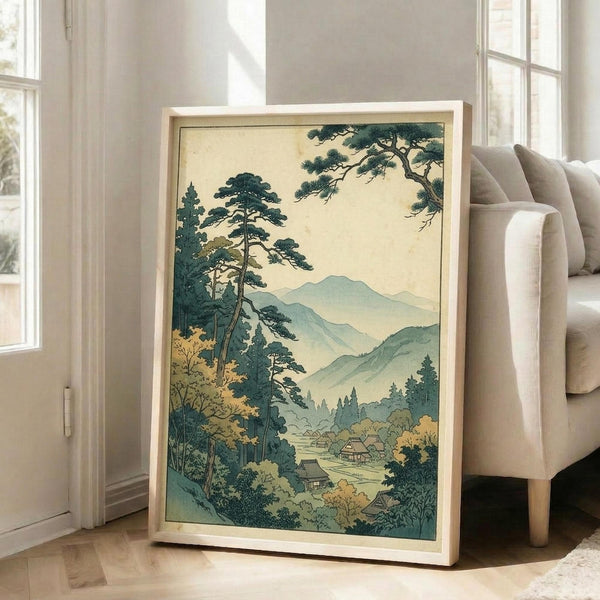 May include: Framed vintage Japanese woodblock print artwork leaning against white wall next to beige upholstered chair. The print features misty mountains, tall pine trees, and traditional houses in a valley, rendered in muted greens, blues, and cream tones. Light wooden frame surrounds the serene landscape scene. Natural sunlight streams through nearby windows onto hardwood flooring.