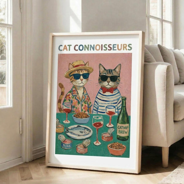 May include: Framed poster print leaning against white wall next to beige sofa in bright living room. Artwork shows two cats wearing sunglasses and clothes sitting at turquoise table with wine glasses, fish, bowls, and green bottle labeled "Catnip Brew." Title reads "Cat Connoisseurs" in colorful letters against pink background. Light wood frame with white matting.