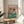 May include: Framed poster print featuring two illustrated cats wearing sunglasses sitting at a turquoise table with food and drinks. One cat wears a straw hat and floral shirt, the other has a striped shirt and red bow tie. The artwork has a pink background with "CAT CONNOISSEURS" text at top. Print is displayed in a thin wooden frame on a round wooden side table next to a beige upholstered chair, with natural sunlight and plant shadows visible.