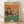 May include: Framed vintage-style poster showing two anthropomorphic cats playing poker at green felt table. Orange tabby cat wears sunglasses, burgundy jacket, smoking cigar. Second cat in pearl necklace holds martini glass. Poker chips, whiskey bottle, playing cards, and small fish scattered on table. Warm orange background. Light wood frame displayed on wooden floor against white wall.