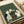 May include: Framed art print on cream paper showing five cats gathered around a wooden basket filled with colorful yarn balls. The illustration features white, black, and striped cats in a vintage woodcut style against a green textured background. The print sits on white fabric beside a rolled paper tube, displaying the artwork's warm earth tones and detailed linework reminiscent of traditional printmaking techniques.