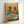 May include: Framed poster print leaning against white wall showing two anthropomorphic pigeons in human clothing sitting at green cafe table. Left pigeon wears denim jacket and beret, right pigeon in tweed suit with bow tie and sunglasses. Table displays avocado toast, pancakes, croissant, coffee cups with latte art, and newspaper. Warm orange painted background, vintage illustration style in light wood frame.