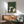 May include: Framed poster showing cats gathered around a small table with yarn balls in Japanese woodblock print style. Green textured background with cats in white, black, and gray colors. Wooden frame hung above mid-century modern walnut credenza with white ceramic lamp. Green upholstered chair with wooden arms sits nearby in sunlit room with white walls.