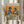 May include: Person in cream sweater holding framed art print against white curtained window, with dried wheat stems visible to left. Print shows two stylishly dressed pigeons with sunglasses sitting at green café table with coffee cups, avocado toast, croissant, and pancakes. Orange painted background with colorful "PIGEON BRUNCH" text at top. Light wood frame, cozy indoor setting with natural lighting.