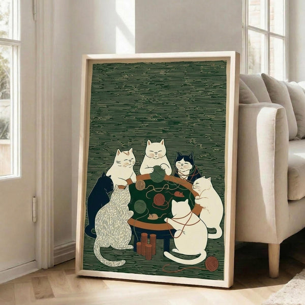 May include: Framed art print in light wood frame showing six cats gathered around a green table with yarn balls and knitting supplies, styled as a parody of classic poker paintings. Green textured background with horizontal line pattern. Print displays in bright living room setting next to beige sectional sofa and large windows with natural lighting on hardwood floors.
