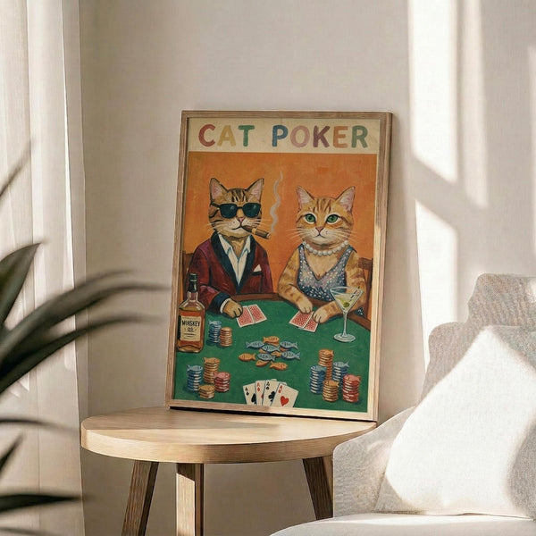 May include: Framed poster displaying two anthropomorphic cats playing poker at a green felt table. One tabby cat wears sunglasses and burgundy jacket while smoking, the other orange cat wears pearls and holds a martini. Colorful poker chips, playing cards, and whiskey bottle scattered on table. Orange background with "CAT POKER" text in rainbow letters at top. Print shown in thin wooden frame on round wooden side table next to beige upholstered chair and plant leaves.