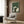 May include: Framed poster print showing cats gathered around a small table with yarn balls in a green textured background, displayed on wood paneled wall above beige upholstered armchair with geometric patterned pillow, brass floor lamp and side table with open book nearby in modern living room setting
