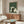 May include: Framed poster print hanging above wooden credenza showing cartoon cats gathered around table with green yarn balls in Japanese woodblock print style with green textured background. Modern living room setting with white curved chair, glass coffee table, floor lamp, and neutral walls.