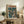 May include: Framed anime-style poster featuring a character in a brown trench coat standing in a rainy urban street scene with teal and turquoise tones and Japanese text at the top, displayed on a rustic wooden sideboard alongside books, a ceramic vase, and fern plant in a cozy living room with beige armchair and warm lighting