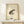 May include: Framed traditional Japanese ink wash artwork featuring a spotted brown and beige frog with detailed eyes, positioned on cream-colored aged paper with red seal stamps and black calligraphy characters in upper left corner. Natural wood frame sits on light wooden shelf next to small white wrapped gift with evergreen sprig against neutral wall.