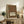 May include: Framed vintage-style poster with ornate floral border in earth tones featuring birds, flowers, and decorative text reading "ART & SOCIALISM" displayed on rustic wooden sideboard alongside fern plant, books, ceramic vase, and brown leather chair in cozy living room setting with cream walls