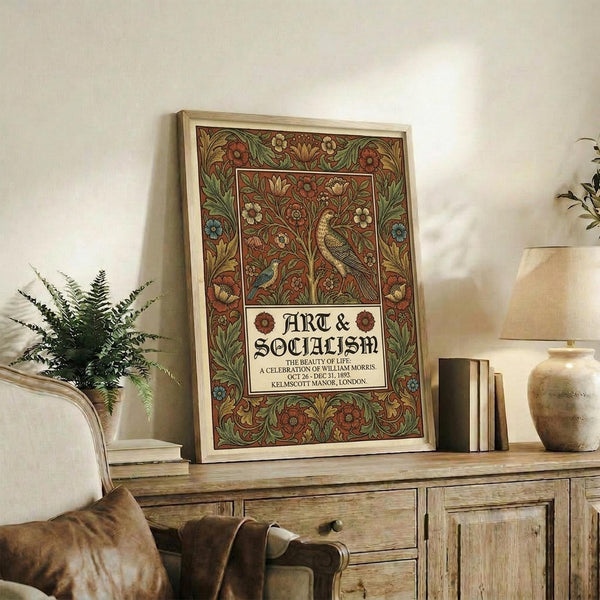 May include: Framed vintage-style poster with ornate floral border in earth tones featuring birds, flowers, and decorative text reading "ART & SOCIALISM" displayed on rustic wooden sideboard alongside fern plant, books, ceramic vase, and brown leather chair in cozy living room setting with cream walls