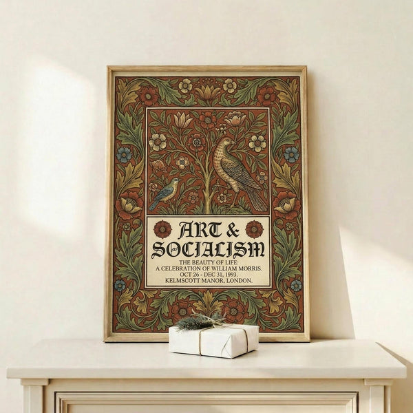 May include: Framed Arts and Crafts style poster featuring ornate floral border with birds, flowers, and foliage in rich burgundy, green, and gold tones. White text panel reads "Art & Socialism" with exhibition details. Natural wood frame displayed on light wooden console with small wrapped gift. Cream colored wall background.