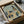 May include: Framed abstract architectural artwork in muted blue, green, orange and beige tones featuring geometric buildings and industrial structures in cubist style, carefully packed in brown cardboard box with protective tissue paper wrapping and wooden corner guards