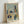 A framed abstract modernist artwork featuring geometric buildings and architectural forms in muted blues, oranges, greens, and beige tones. The composition includes simplified trees, angular structures, and overlapping geometric shapes creating a cubist-style cityscape. The piece has a soft watercolor-like background with defined linear elements throughout. The artwork is displayed in a natural wood frame, leaning against a white wall.