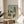 May include: Abstract modernist artwork in wooden frame displayed on round wooden side table next to beige upholstered chair. The print features geometric buildings in blue, orange, yellow and green watercolor tones with stylized trees against a soft turquoise sky. Natural sunlight streams across the neutral-toned room setting with plant shadows visible.