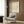 May include: Minimalist line drawing of a bull in black on cream background, framed poster on wood-paneled wall above beige mid-century modern armchair with geometric patterned pillow, gold floor lamp, and dark round side table with open book in contemporary neutral-toned room